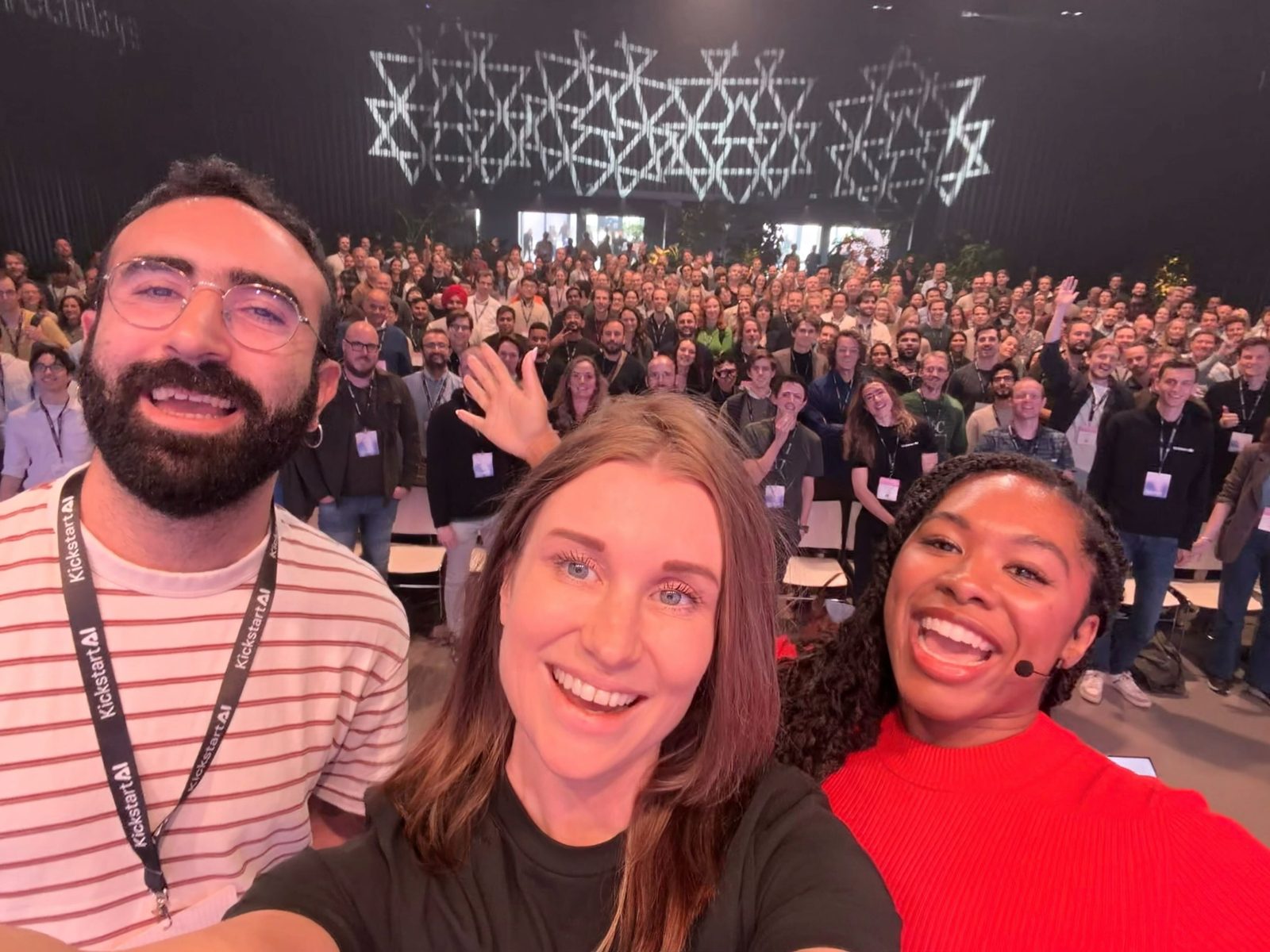 A group selfie taken from the main stage: three people smiling in front, and a full hall of attendees with lanyards waving behind them.