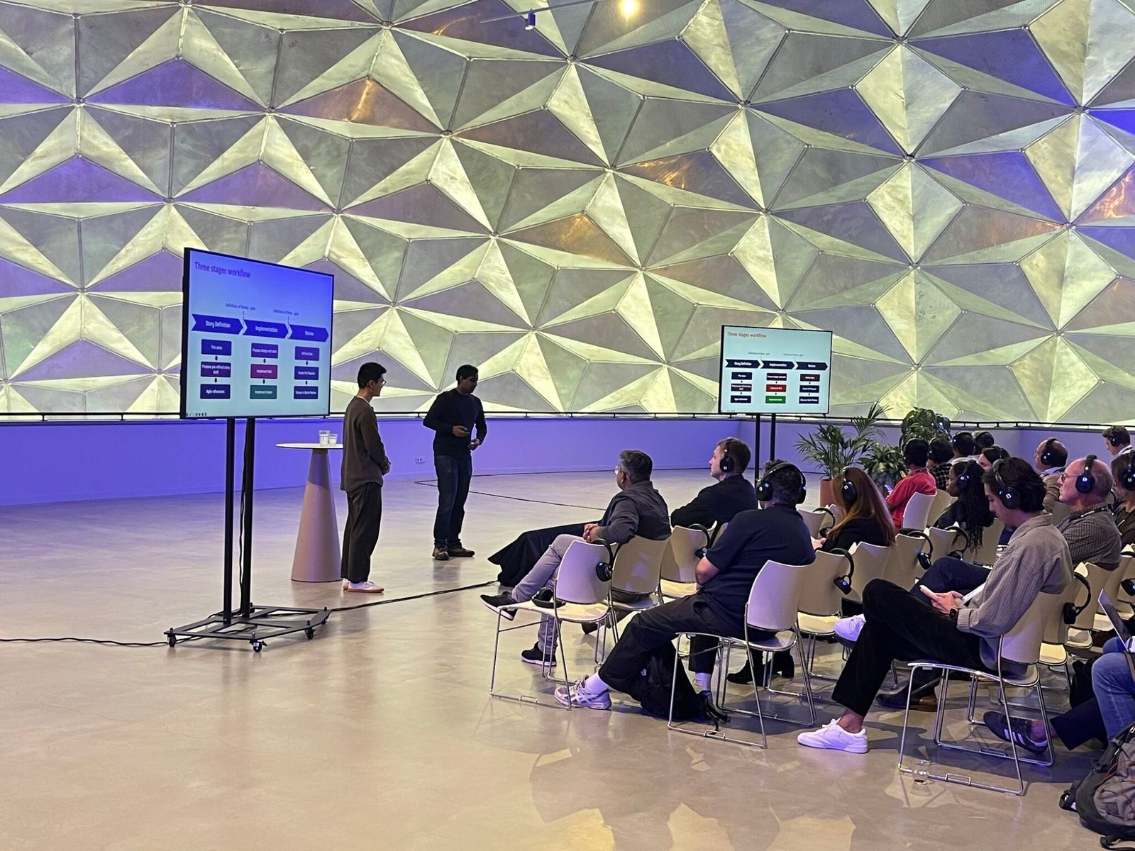 Two speakers stand next to a screen showing a three stage workflow slide, under a faceted metal dome, while an audience wearing headphones watches from rows of chairs.